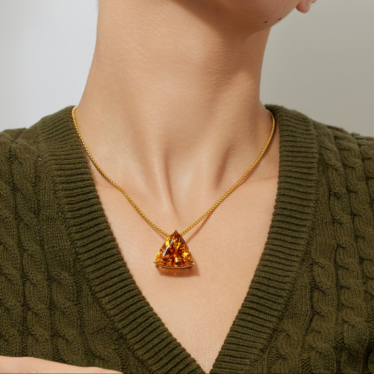 Woman wearing a gold chain necklace featuring a large trillion-shaped golden orange citrin pendant against a plain background