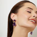 Woman wearing statement bold dark amethyst purple teardrop gold earrings with a plain background